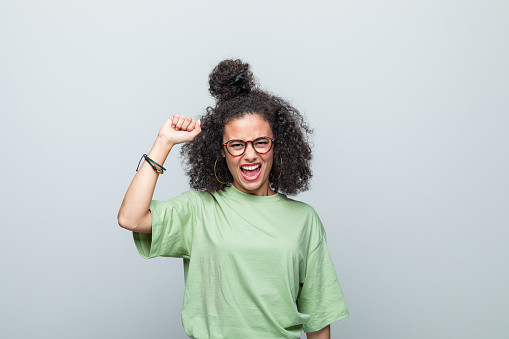 Angry young woman wearing green t-shirt and eyeglasses shouting at camera and raising fist. Studio shot against grey background.