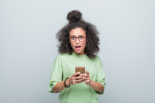 Terrified young woman wearing green t-shirt and eyeglasses holding smart phone in hands and looking at camera. Studio shot against grey background.