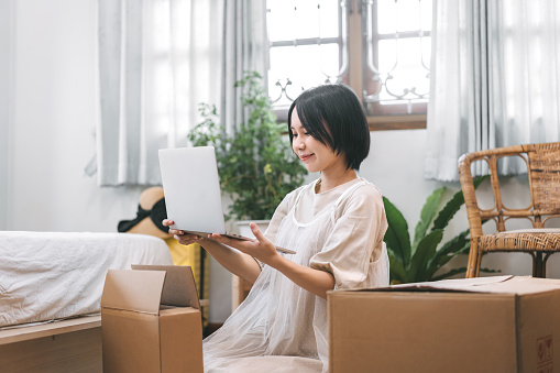 People moving house to new home or apartment concept. Asian woman unpack parcel box for new laptop in bedroom. Background cozy style room with window and carton for relocation.