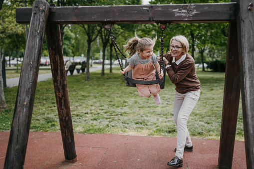 Young grandmother pushing her little granddaughter who is sitting on a swing in public park outdoors.