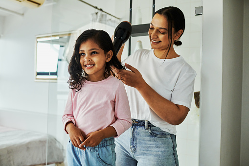 Mother and daughter smiling while blow drying hair in bathroom at home .