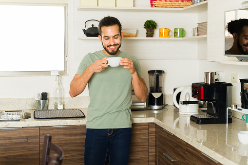 Cheerful young man smiling while enjoying drinking a coffee cup in the kitchen and getting ready to do housework at home