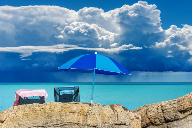 Blue beach umbrella and two deck chairs on the rocky coast with beautiful seascape with storm clouds (cumulonimbus) and torrential rain on background. Gulf of La Spezia, Lerici, Italy, Europe.