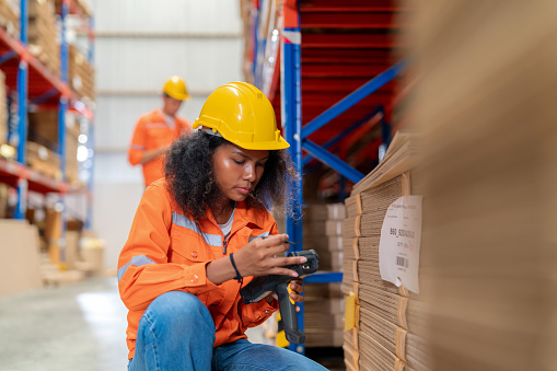 Dedicated warehouse worker overseeing the organization of a factory warehouse. Wearing in reflective protective workwear scanning the barcodes on carton boxes using a modern barcode reader. Ensuring precise identification and tracking of merchandise.
