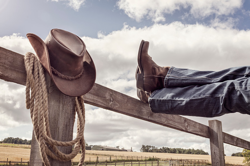 Cowboy boots and hat with feet up on stables fence at ranch resting with legs crossed, country music festival live concert or line dancing concept