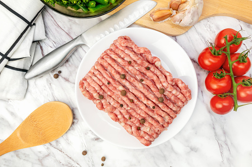 Uncooked gound pork, beef. Fresh raw minced meat with peppercorns on white plate. Ingredients for cooking meal, tomatoes, broccoli, garlic on marble table on kitchen. Top view.