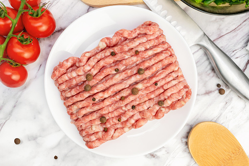 Top view of fresh raw minced meat with peppercorns on plate. Ingredients for cooking meal, tomatoes, broccoli, garlic on marble table. Uncooked gound pork, beef.