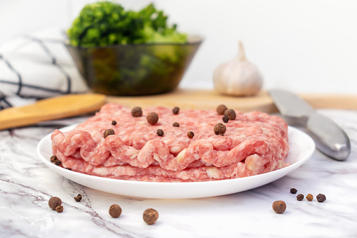 Uncooked gound pork, beef. Fresh raw minced meat with peppercorns on white plate. Ingredients for cooking meal, tomatoes, broccoli, garlic on marble table on kitchen.