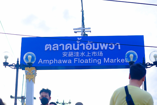 Amphawa Floating market sign and people on bridge captured from steps in low angle view in early evening. People are taking photos with sign and from bridge. Floating market is popular weekend market and travel destination