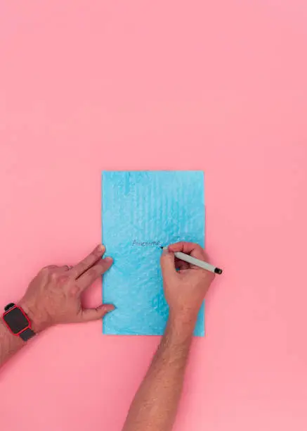 Overhead view of caucasian adult male hands writing the word Awesome on a blue bubble packing envelope. Pink background with room for copy space.