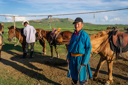 Bulgan Province, Mongolia - July 16, 2023: A nomadic man stands in front of horses tethered to a rope, as another tends to them.