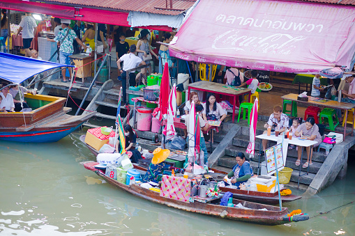 People and tourists are sitting and eating at floating market kitchen in Amphawa. People are sitting on stools on steps at river. Behind is promenade with old houses, restaurants and shops. On river a thai woman is preparing food in  a boat