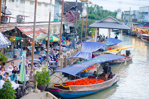 Anchored boats and people at restaurants of floating market in Amphawa. People and tourists are sitting at tables of restaurant on promenade.  Scene is at river and floating market. View from one of several bridges between promenades of both sides.