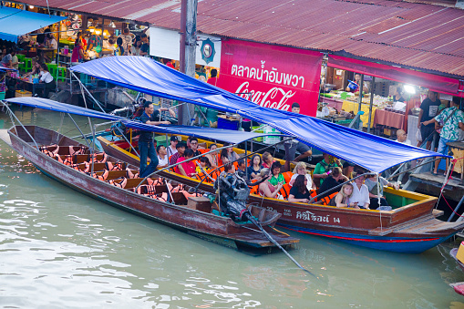 Asian and  white people tourists in tourboat in Amphawa in floating market area. View from one of bridges between promenades at both sides of river. People are wearing life jackets. Behind boat people are at a restaurant and promenade.