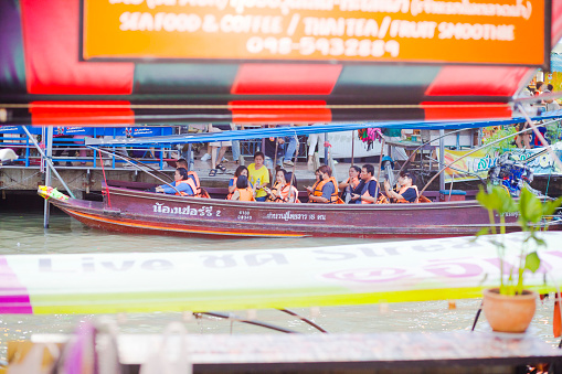 Group of tourists in tourboat in Amphawa captured on almost water level of river.  Boat is anchored in old part of Amphawa with floating market area and many shops and restaurants in old wooden houses. Tourboats are making trips along Mae Khlong river around Amphawa. People are wearing life jackets