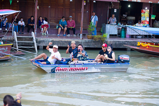 Thai people on promenade and in small boat in Amphawa. Scene is in area of floating market with old wooden houses. A group of men is sitting inside of a small boat. In background people are in front of houses and restaurants