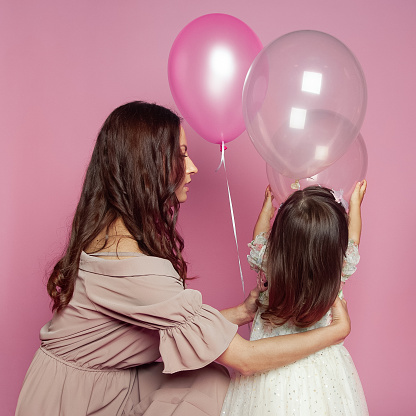 Brunette mother and daughter with ballon on pink studio background. Mom and cute child baby girl