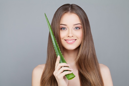 Cheerful woman with long straight hair, shiny clear skin and cute smile showing natural green aloe vera leaf. Facial treatment, medicine and skincare concept