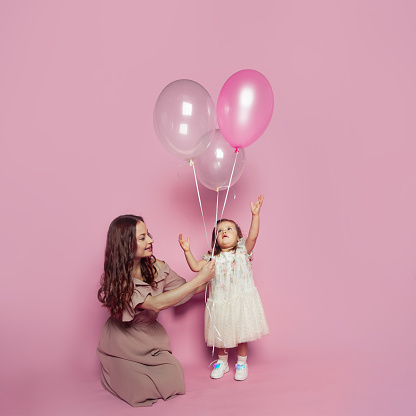 Cheerful adorable mother and daughter with pink ballon on pink studio background. Portrait of young mom and cute child baby girl