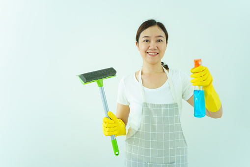 Happy beautiful Asian woman or female housewife posing to camera with showing a rubber squeegee and liquid glass cleaning solution on white wall background.