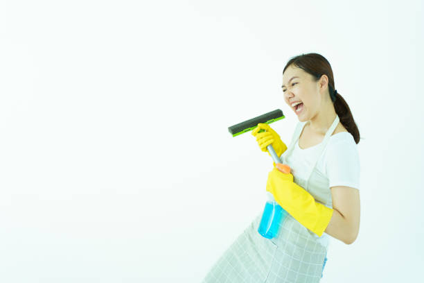 Happy beautiful Asian woman or female housewife posing to camera with showing a rubber squeegee and liquid glass cleaning solution on white wall background.