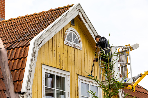 Enkoping, Sweden July 12, 2023 A person on a ladder scraping and preparing a yellow house for painting.