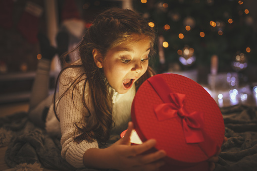 A surprised little girl is lying down on the floor beside Christmas tree and looking at the gift in the red box she just received.