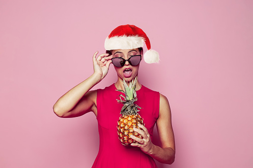 Christmas woman Santa holding pineapple tropical fruit isolated on pink background.Female model in Santa hat, Christmas and New Year holiday and vacation concept