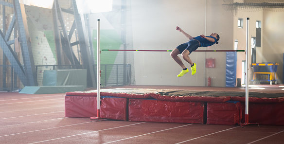 Sportswoman high jumping through pole and falling on exercise mat while training in gym wide shot