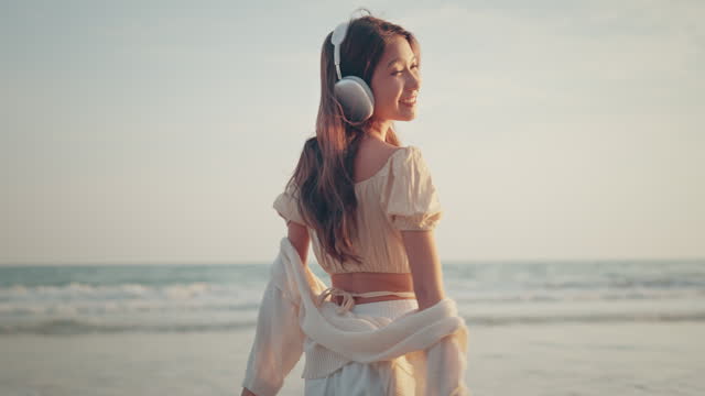 Relax asian woman wearing headphones walking in the sea beach at golden sunset - Female tourist listening to music on summer vacation
