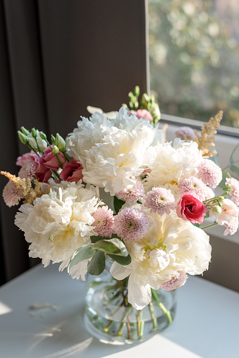 An elaborate bouquet of flowers in  a vase.  All the flowers are white with some greenery mixed in.  The vase sits on a marble surface and in front of a blank gray background.  The lighting is natural and soft.  Nobody is in the photograph.