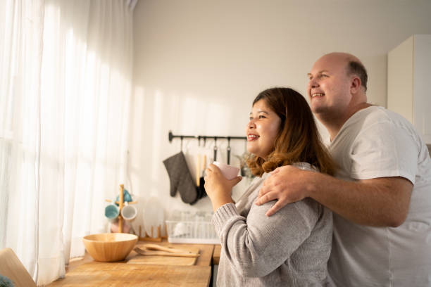 Love Blossoms at Home: Newlyweds Share a Tender Hug and Proposal Witness the heartwarming scene as a newly married couple embraces in their home. Amidst the comfort of their space, a heartfelt proposal seals their love with a promise for a beautiful future together. fat asian couple stock pictures, royalty-free photos & images