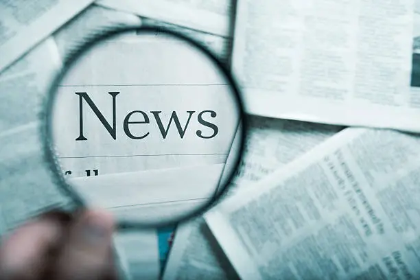 man holding a magnifying glass on the "News" word in the stack of newspapers, blue toned image