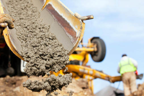 A construction worker is guiding a cement mixer truck chute as concrete is being offloaded for a new house footing. The background is overcast blue sky with another worker, wearing a safety vest, cleaning a mixer chute.\u2028http://www.banksphotos.com/LightboxBanners/Concrete.jpg