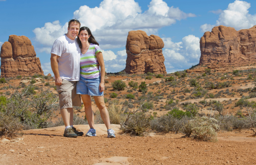 Typical tourist photo: Middle aged man and woman with arms around each other smile for the camera with tall rock columns of Arches National Park in the background. Puffy clouds fill a deep blue sky.