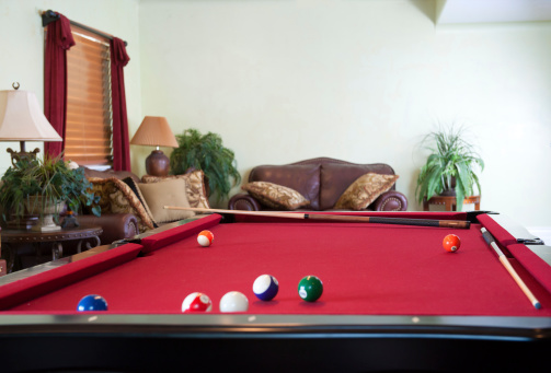 Home interior: Pool balls and cue sticks lay on a red felt pool table in a home interior scene depicting a game room.
