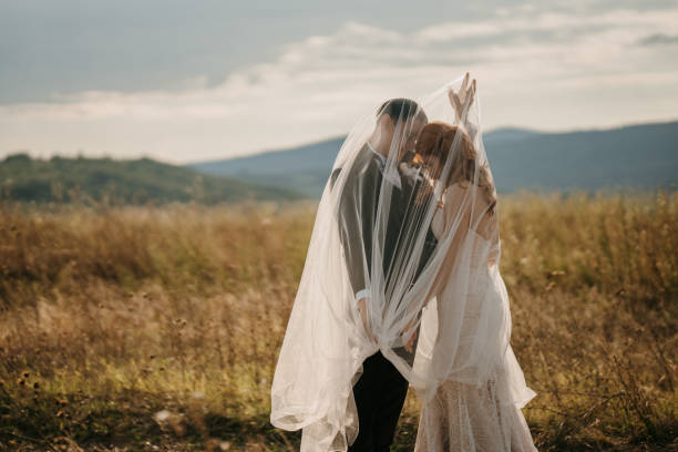 Just married young couple standing under long veil while holding hands in nature