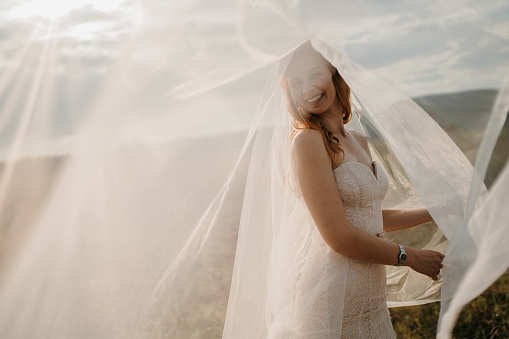 Happy young bride standing under a long veil in nature