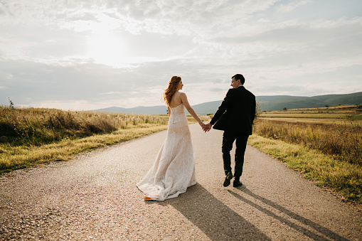 Back view of happy bride and groom holding hands while walking on empty road in nature