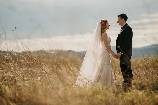 Full length shot of just married young couple standing and holding hands while looking at each other in nature