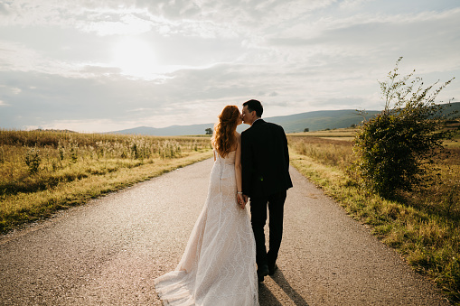 Back view of happy bride and groom holding hands and kissing while walking on empty road in nature