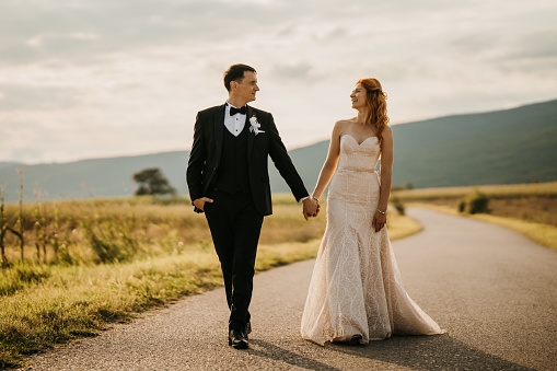 Happy bride and groom holding hands while walking forward on empty road in nature