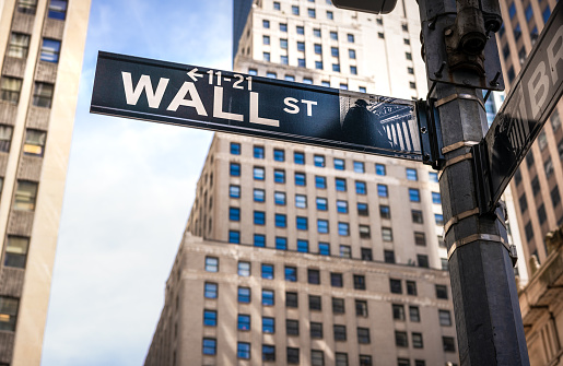 Close-up on a sign for Wall Street, with Lower Manhattan's office skyscrapers in the background.