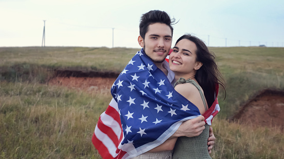 Young couple kiss tenderly wrapped in American flag on hilly field in cloudy day. Concept of holiday of country and people in love