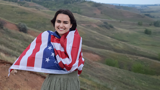 Cheerful female wraps in USA flag standing on hilly place with picturesque view. Concept of love for country and countryside