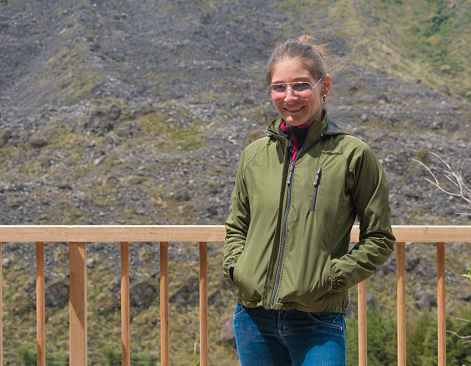 Young explorer woman hiking, smiling at camera on a wooden deck with rocky mountain on the background. Freedom and adventure concept.
