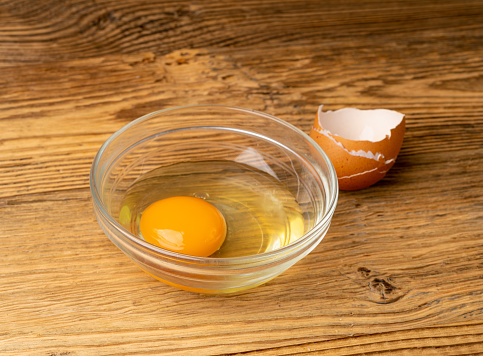 One Broken Egg in Glass Bowl, Raw Yolk and White, Fresh Broken Organic Chicken Egg on Wooden Rustic Background