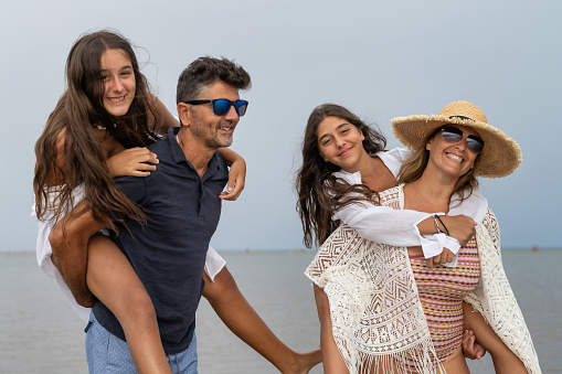 Happy Family Having Fun Outdoors. Parents Giving Piggyback Rides To Children On The Beach. Happy Couple With Their Children Smiling.