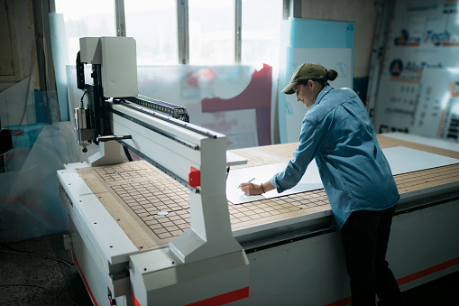 Woman marking on workpiece over cnc router machine flatbed in signboard making factory. Female manual worker working at signable manufacturing company.