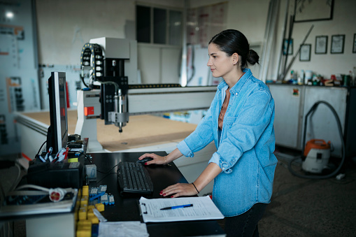 Side view of a female programming a cnc router machine for making acrylic signs in workshop. Young caucasian woman making a computer program for CNC machine at sign board making factory.